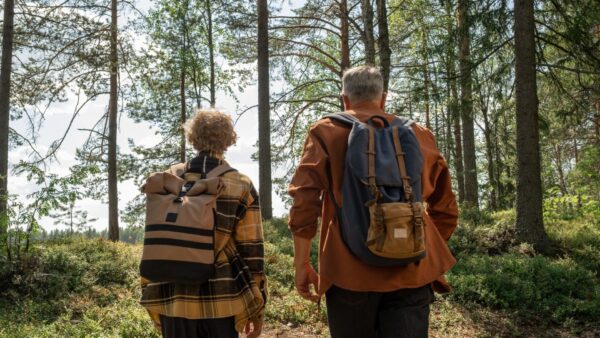 Harmaahiuksinen mies ja kiharatukkainen nuori kävelevät reput selässä mäntymetsän polulla